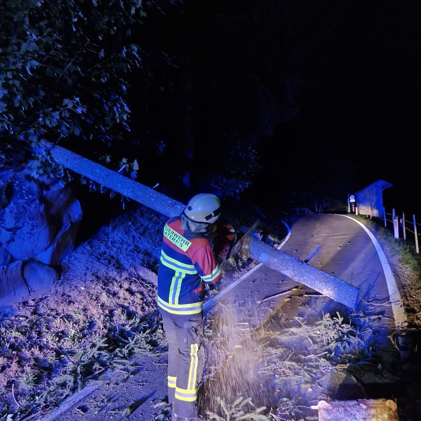 Ein Feuerwehrmann in voller Ausrüstung schneidet einen umgestürzten Ast mit einer Kettensäge in der dunklen Nacht.