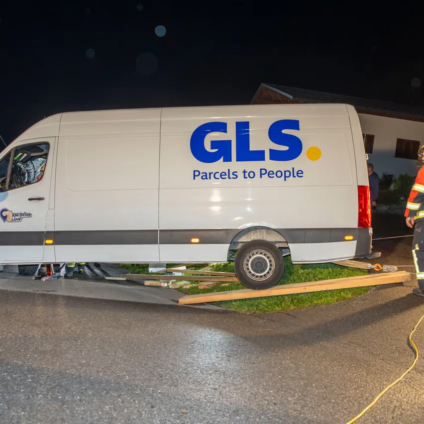 Ein weißer GLS-Van ist nachts auf der Seite einer Straße umgekippt. Ein Feuerwehrmann steht in der Nähe, und ein Haus ist im Hintergrund zu sehen.