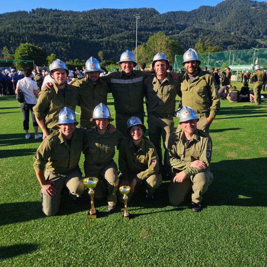 Eine Gruppe von Soldaten in Uniform posiert mit Trophäen auf einem Feld. Dahinter beobachtet eine Menge von den Seitenlinien.