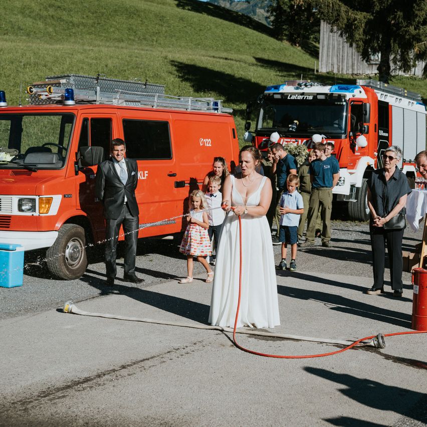 Eine Hochzeitszeremonie findet im Freien statt, mit einem Feuerwehrauto im Hintergrund. Die Braut und der Bräutigam stehen vor einem Feuerhydranten, wobei die Braut einen Schlauch hält.