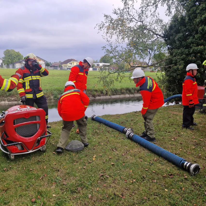 Eine Gruppe von Feuerwehrleuten versammelt sich um ein rotes Fahrzeug, möglicherweise ein Feuerwehrauto, auf einem Rasen in der Nähe eines Gewässers. Sie tragen rote Uniformen und Helme.