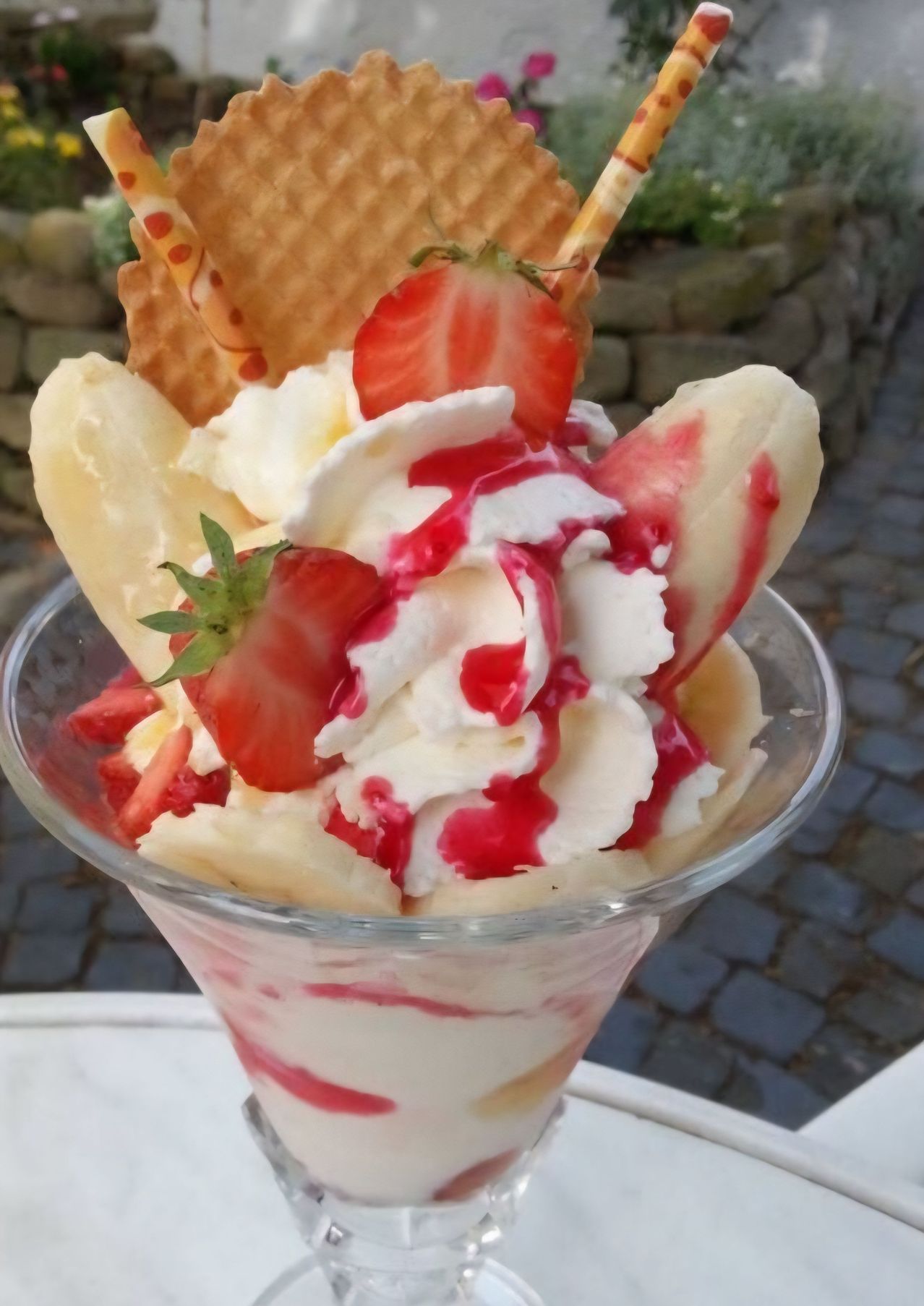 A sundae made with bananas, whipped cream, strawberries, and red syrup. It's in a glass cup with a stone wall in the background.