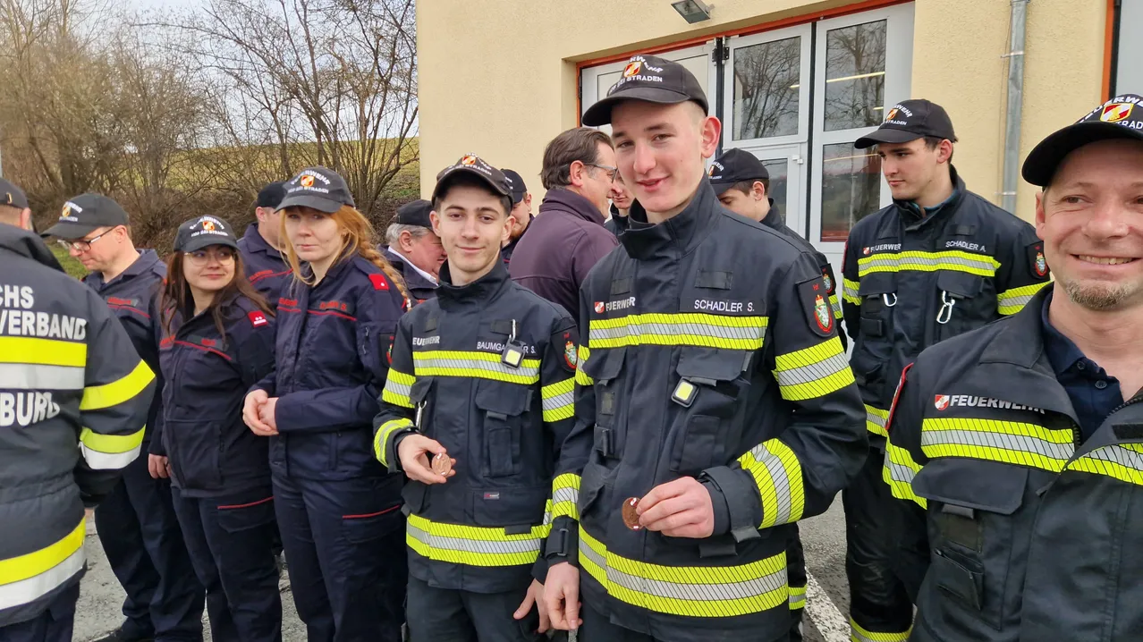 Eine Gruppe Feuerwehrleute, darunter zwei junge Männer, stehen vor einem Gebäude. Sie tragen Uniformen und Mützen mit Abzeichen. Ein Mann hält eine Münze.