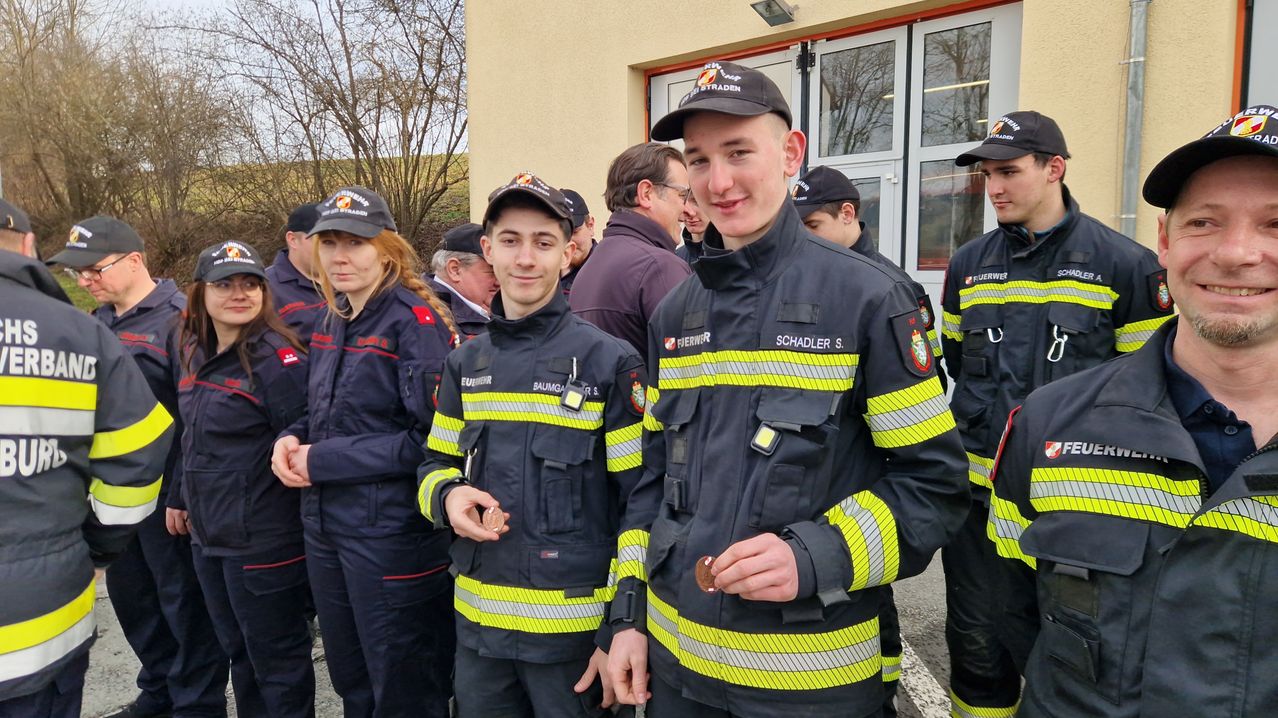 A group of firefighters, including two young men, stand outside a building. They are wearing uniforms and caps with badges. One man holds a coin.