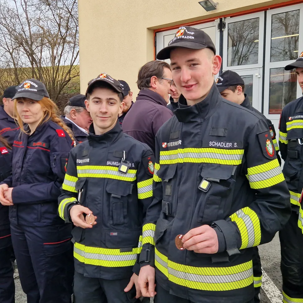 Eine Gruppe Feuerwehrleute, darunter zwei junge Männer, stehen vor einem Gebäude. Sie tragen Uniformen und Mützen mit Abzeichen. Ein Mann hält eine Münze.