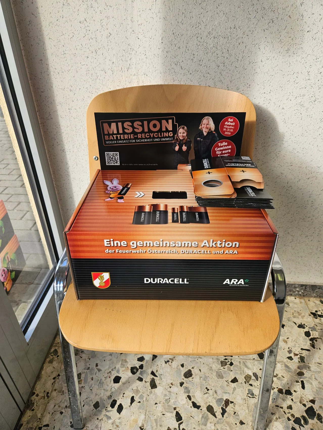Ein Holzstuhl mit einer Schachtel darauf. Die Schachtel hat ein Etikett, das 'Mission Batterie-Recycling' und einen QR-Code liest.