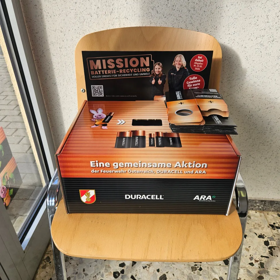 Ein Holzstuhl mit einer Schachtel darauf. Die Schachtel hat ein Etikett, das 'Mission Batterie-Recycling' und einen QR-Code liest.