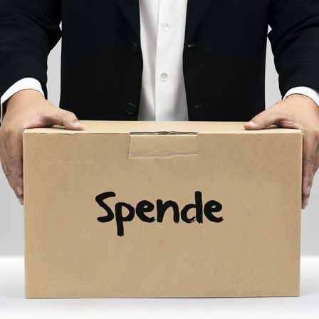 Eine Person in formeller Kleidung hält eine Pappschachtel mit dem Wort 'Spende' darauf, was auf den Akt des Spendens oder Gebens hinweist.