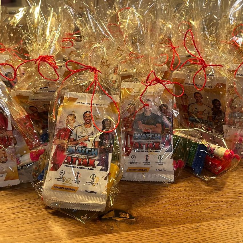 Mehrere Packungen eines Sammelkartenspiels sind in Plastik verpackt und mit roten Bändern gebunden, auf einem Holztisch ausgestellt.