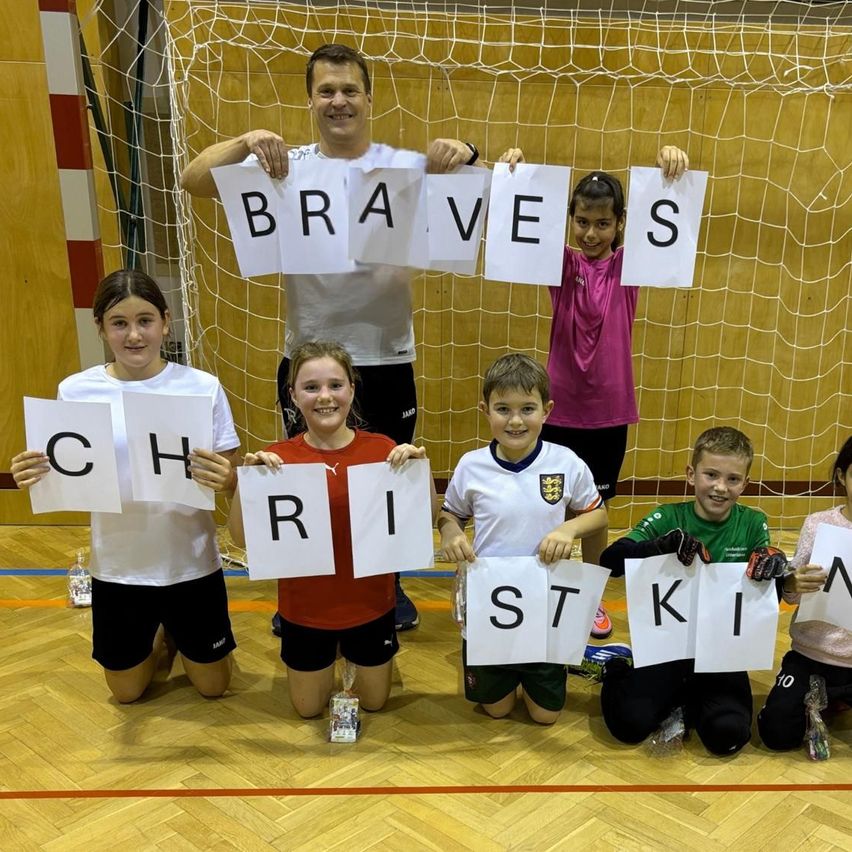 Mehrere Kinder halten Schilder, die 'CHRISTIAN' buchstabieren, während sie vor einem Tor knien. Dahinter steht ein lächelnder erwachsener Mann. Sie befinden sich in einer Sporthalle.
