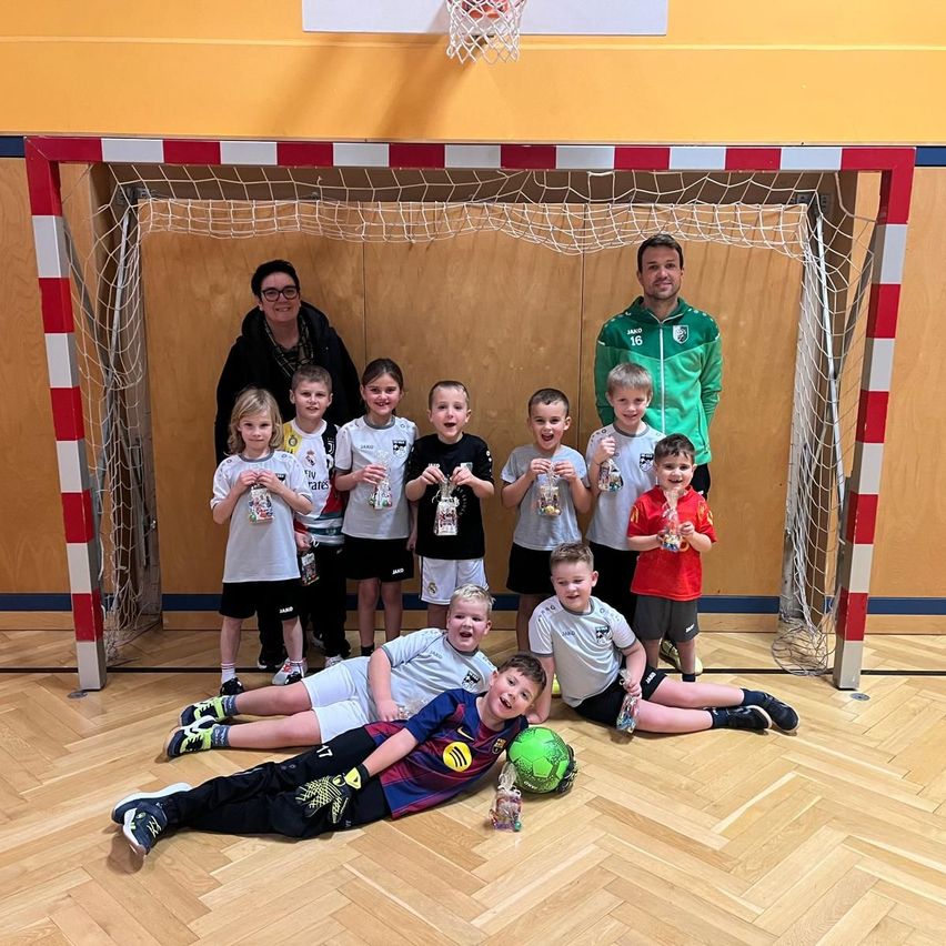 Eine Gruppe von Kindern mit Medaillen und einem Fußball posiert vor einem Tor mit zwei Erwachsenen, einer in einem grünen und der andere in einem schwarzen Jackett.