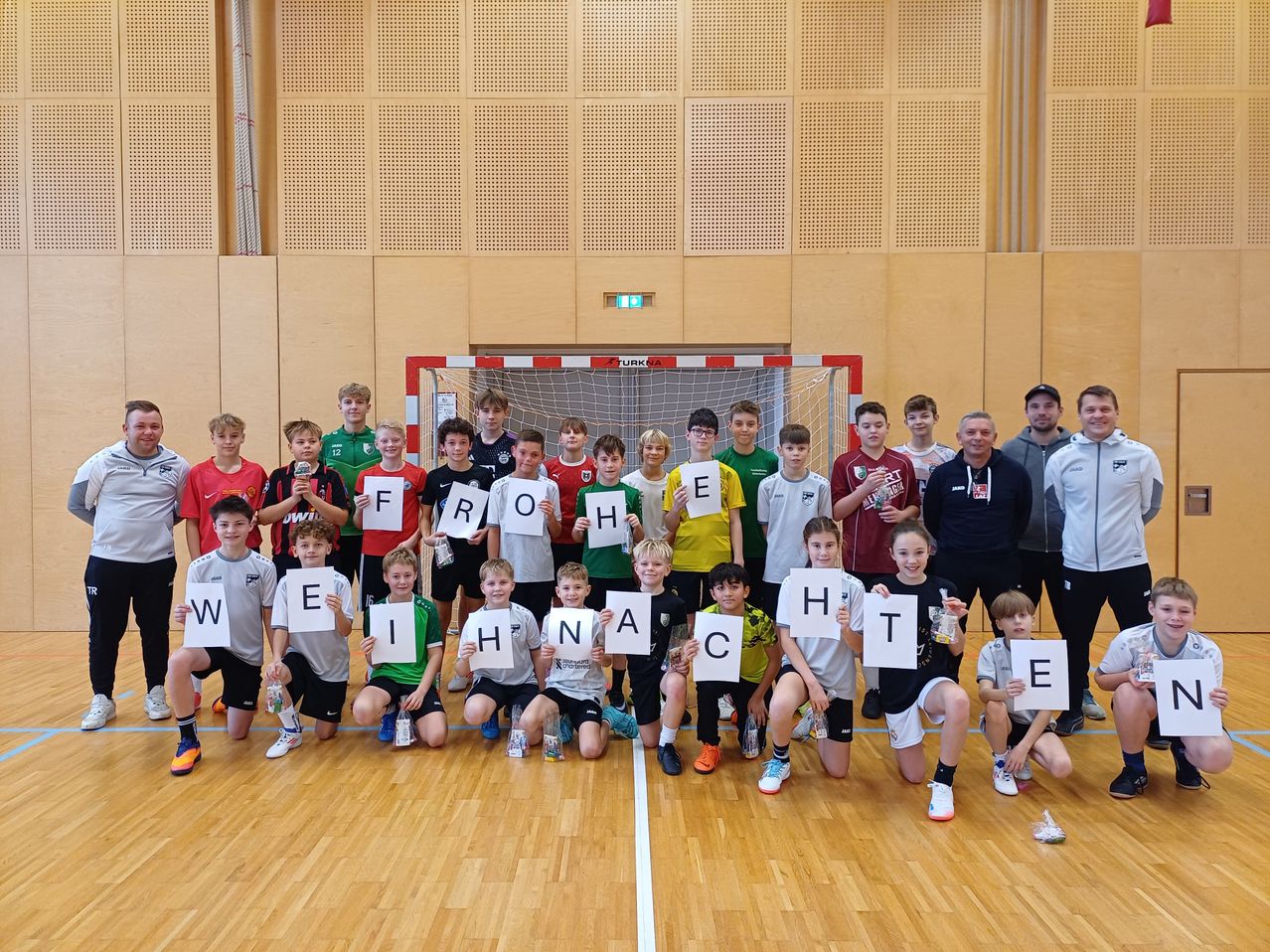 Eine Gruppe junger Athleten, die Buchstabenschilder mit der Aufschrift 'WELKOMEN' halten, posiert vor einem Handballtor.