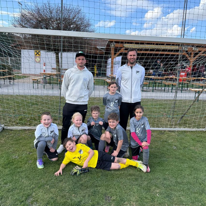 Eine Gruppe von Kindern und zwei Trainern posiert für ein Foto auf einem Fußballfeld mit einem Netz im Hintergrund.
