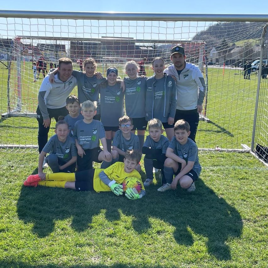 Eine Jugendfußballmannschaft posiert vor einem Tornetz mit zwei Trainern. Die Spieler tragen graue Trikots, einige mit grünen Akzenten. Der Junge in Gelb liegt auf dem Boden und lächelt.