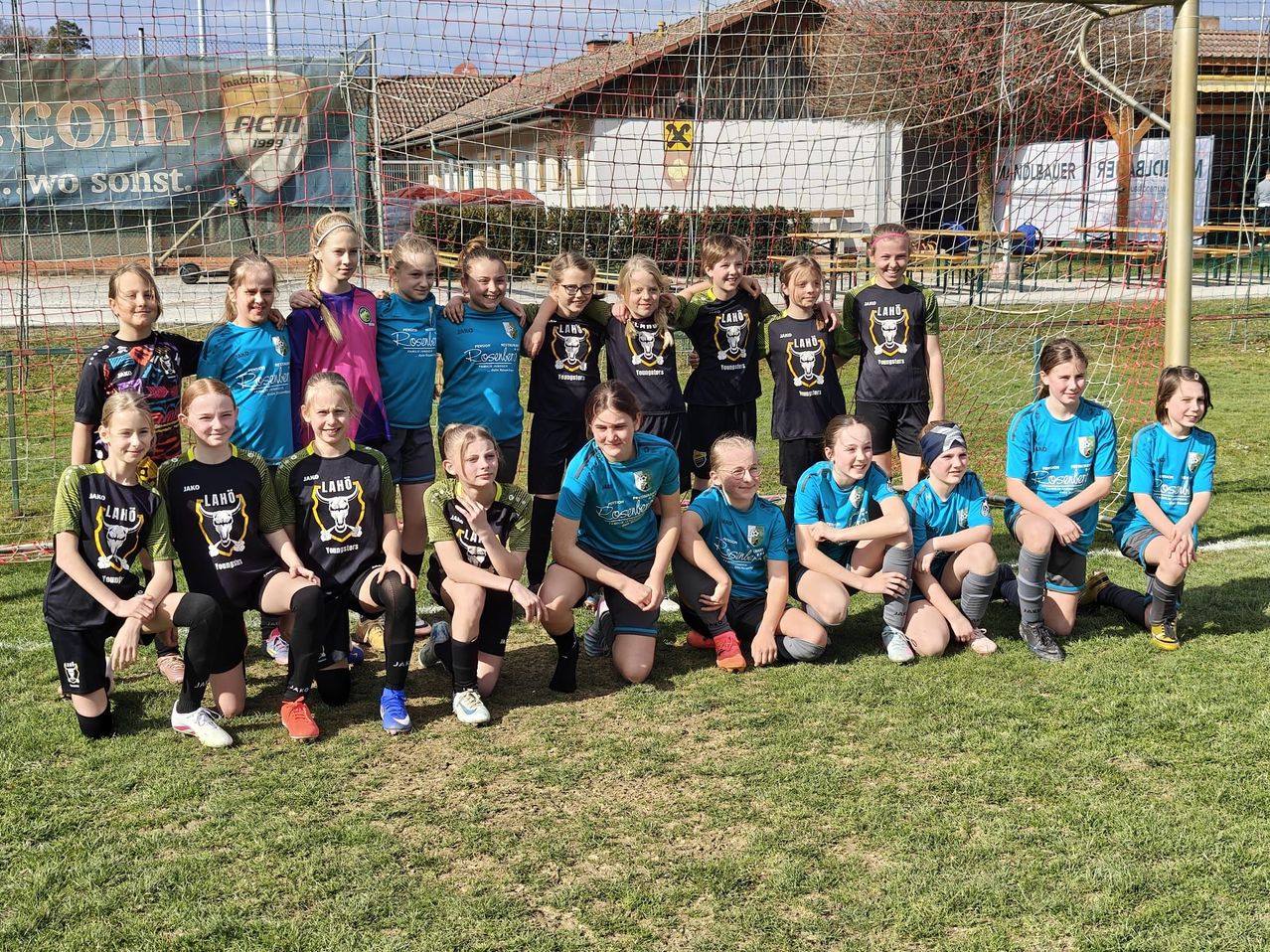 Eine Fußballmannschaft junger Mädchen posiert für ein Foto auf einem Rasen mit einem Netz im Hintergrund.