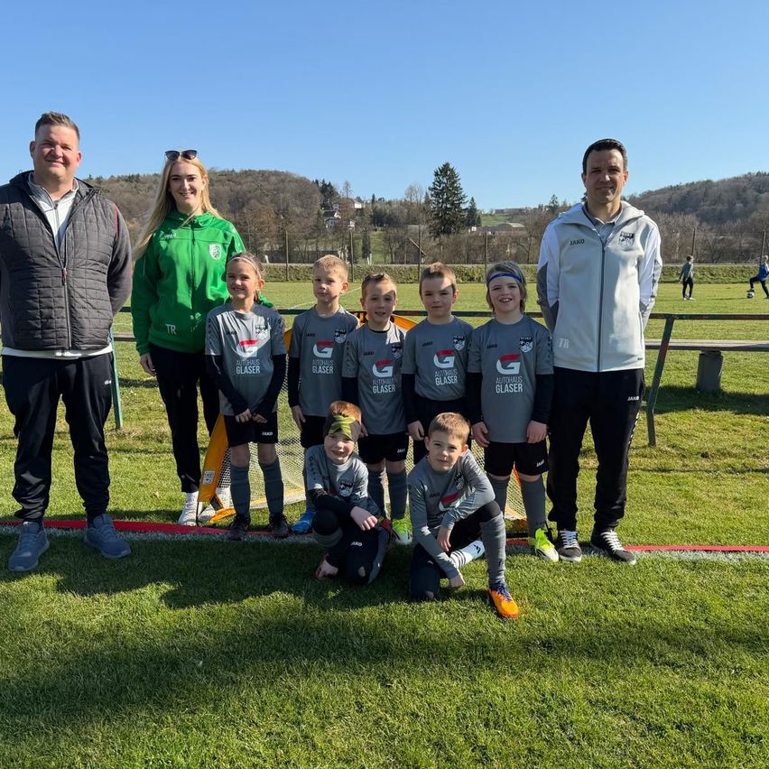 Eine Gruppe von Menschen, einschließlich Kinder und Erwachsene, steht auf einem Fußballfeld und trägt identische Trikots. Sie posieren für ein Foto mit einer lächelnden Frau und zwei Männern im Hintergrund.