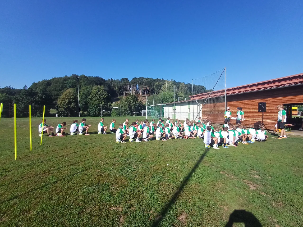 Eine Gruppe junger Athleten in grünen und weißen Uniformen versammelt sich auf einem Rasenfeld, wahrscheinlich in Vorbereitung auf eine Sportveranstaltung. Ein Trainer steht in der Nähe, und ein Gebäude mit einem roten Dach ist im Hintergrund zu sehen.