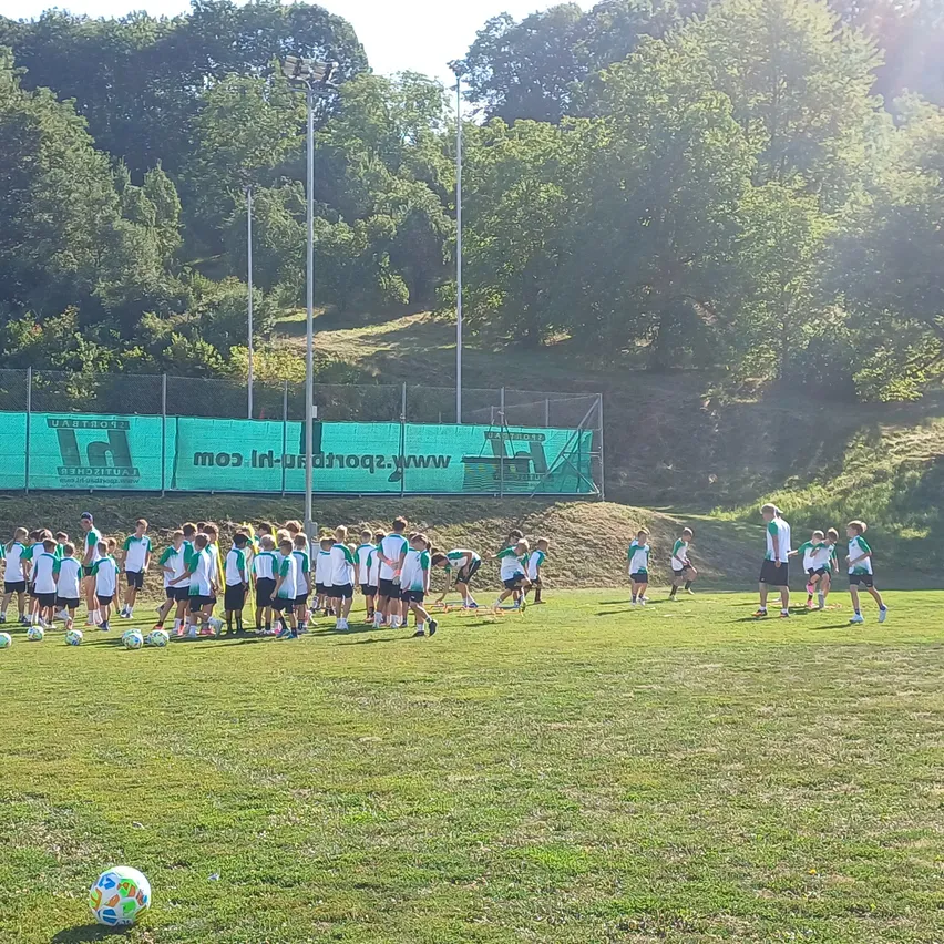 Eine Gruppe junger Fußballspieler in grünen und weißen Trikots trainiert auf einem Rasenfeld mit einem großen grünen Banner im Hintergrund.