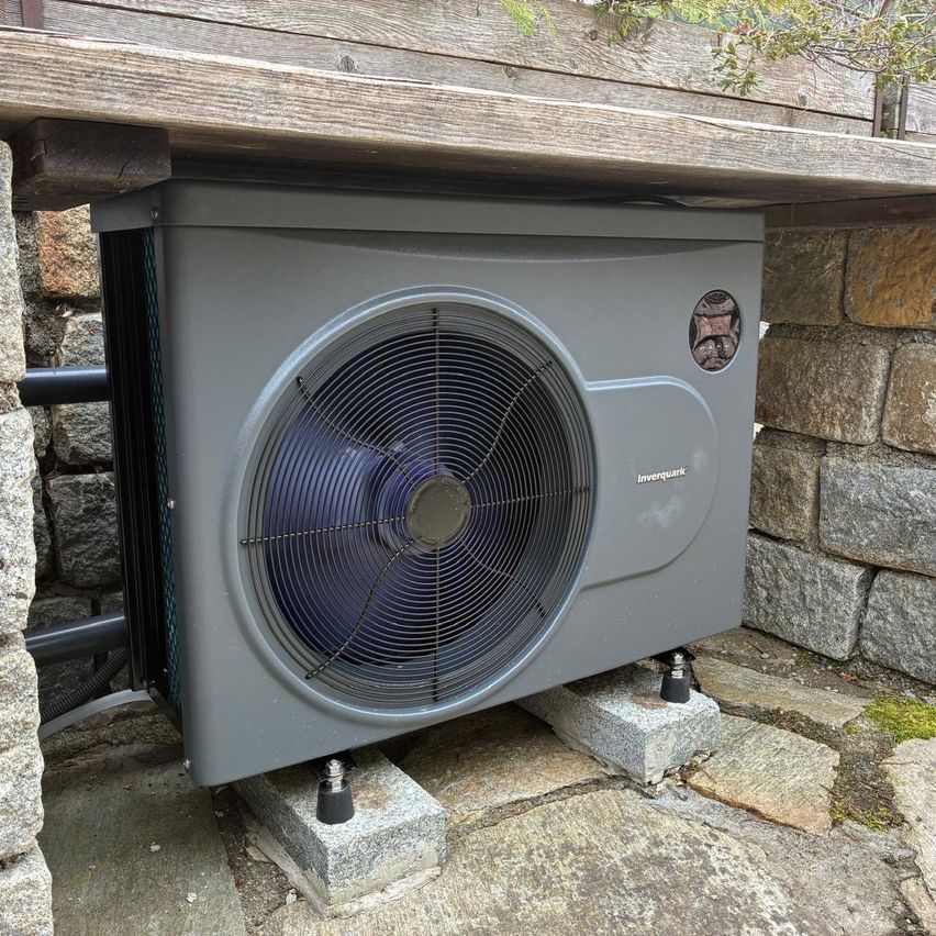 Ein graues Klimaanlagengerät ist unter einer Holzterrasse installiert. Das Gerät hat einen Lüfter an der Vorderseite und wird von Betonblöcken gestützt.