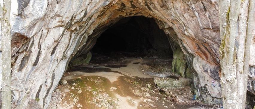 Ein dunkler Höhleneingang mit sandigem Boden und moosbewachsenen Felsen an den Seiten.