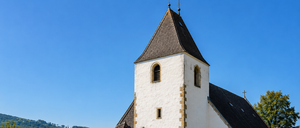 A church with a pointed tower stands near a graveyard. The church has a cross on top and a mural on the side. The graveyard has many tombstones.