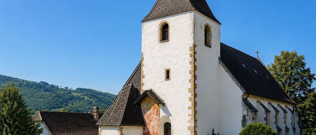Eine weiße Kirche mit einem braunen Dach und einem Turm steht in einem ländlichen Gebiet, umgeben von einem Friedhof mit verschiedenen Grabsteinen und einem Garten mit bunten Blumen.