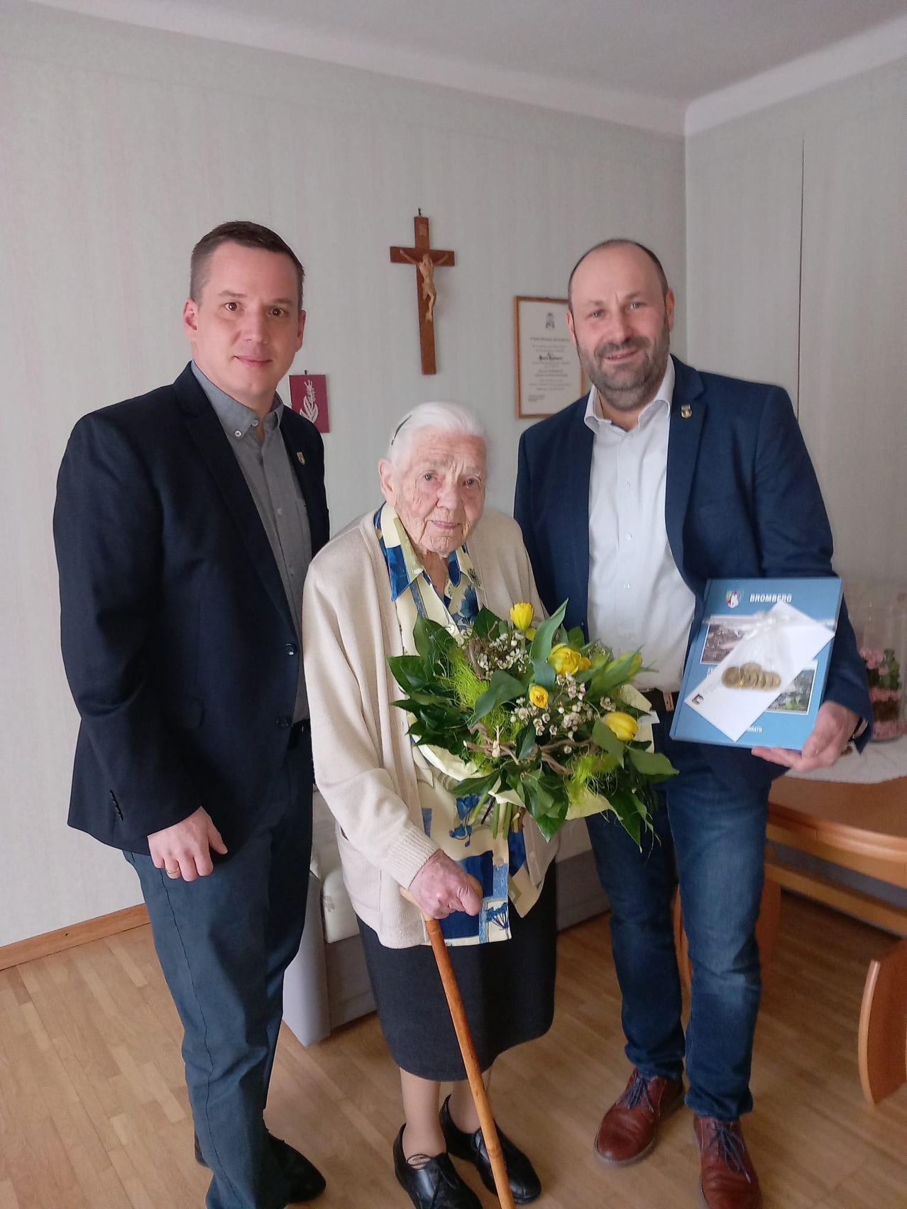 Three individuals stand together in a room; one elderly woman holds a bouquet, and two men hold a folder and a blue book. A cross is mounted on the wall behind them.