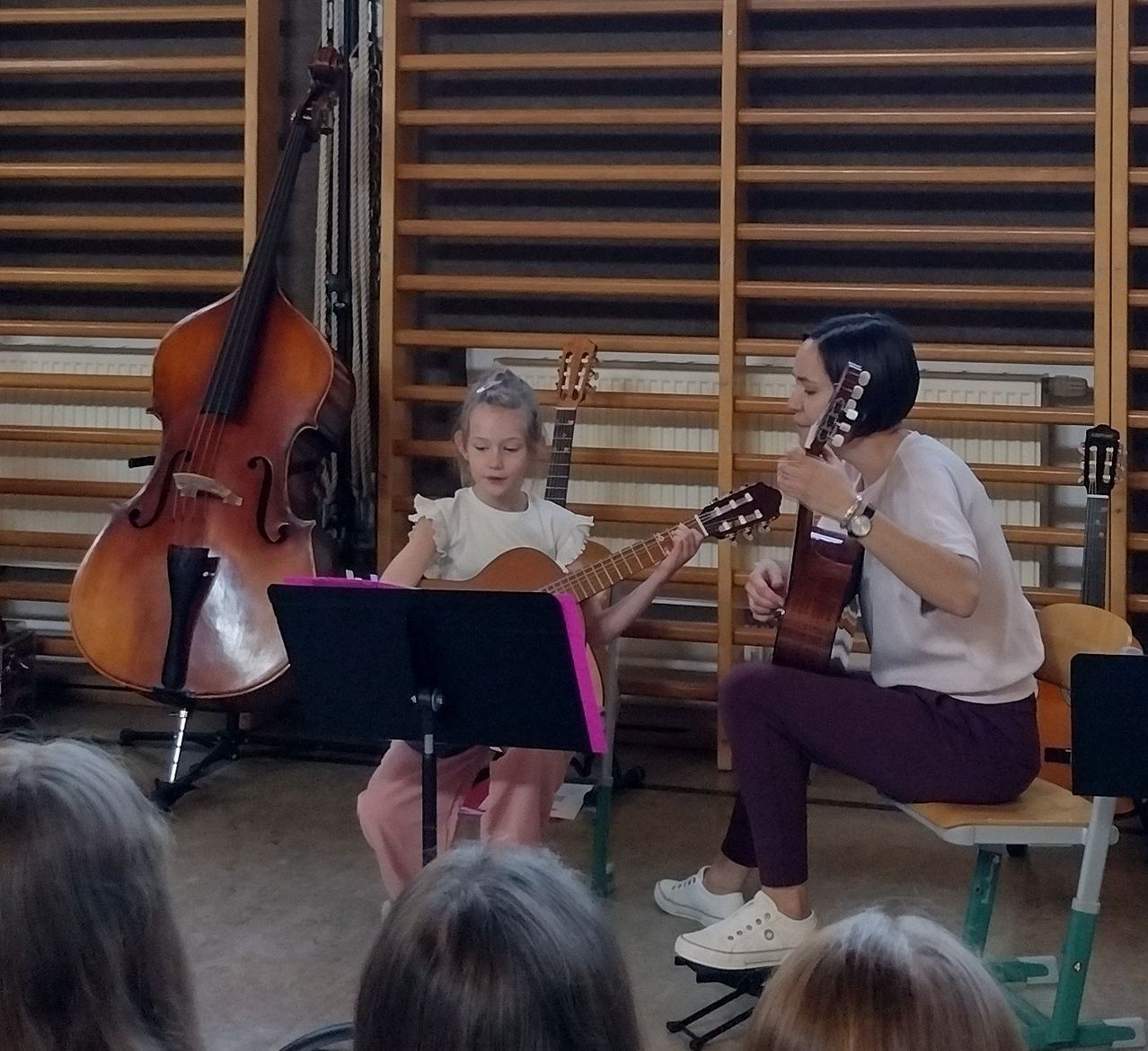 Ein junges Mädchen spielt Gitarre, während eine Frau mit einer Uhr zuschaut. Dahinter steht ein Cello und ein Notenständer. Einige Leute beobachten.
