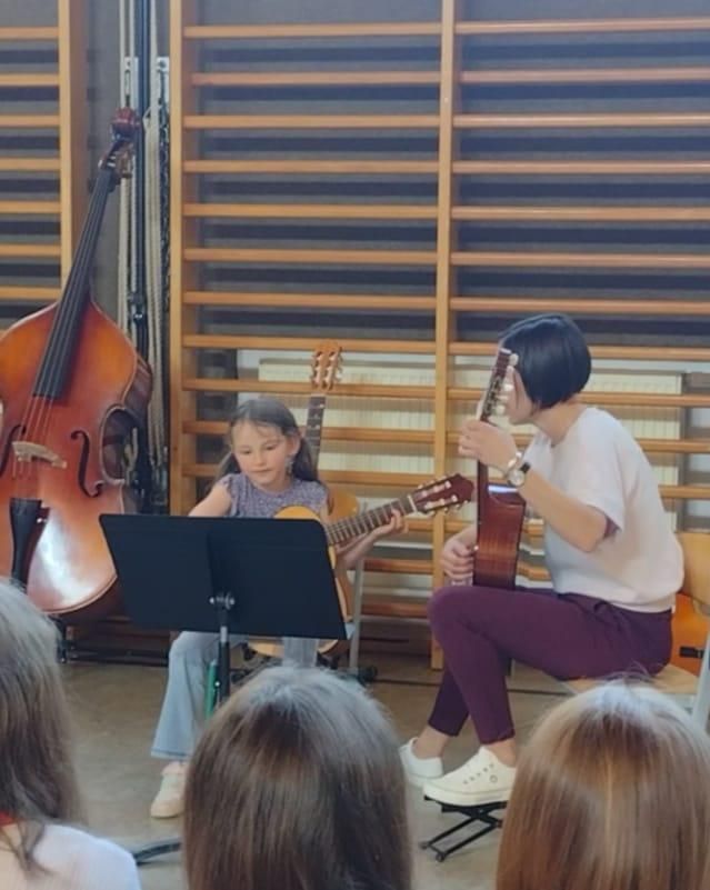 Zwei Mädchen spielen Gitarre in einem Klassenzimmer. Ein Mädchen sitzt auf einem Stuhl und schaut auf ihren Notenständer. Das andere Mädchen sitzt vor ihr und spielt Gitarre. Ein Cello steht hinter ihnen.
