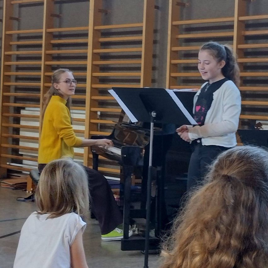 Zwei Mädchen sind in einem Musikraum. Eine spielt Klavier, während die andere neben ihr steht. Ein Publikum beobachtet von vorne.
