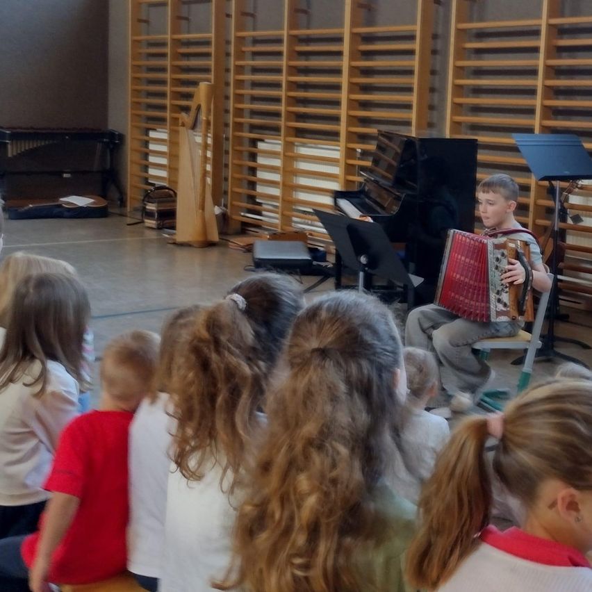 Ein junger Junge spielt Akkordeon für ein Publikum aus Kindern, die in einem Klassenzimmer sitzen. Dahinter sind ein Klavier und eine Harfe zu sehen.