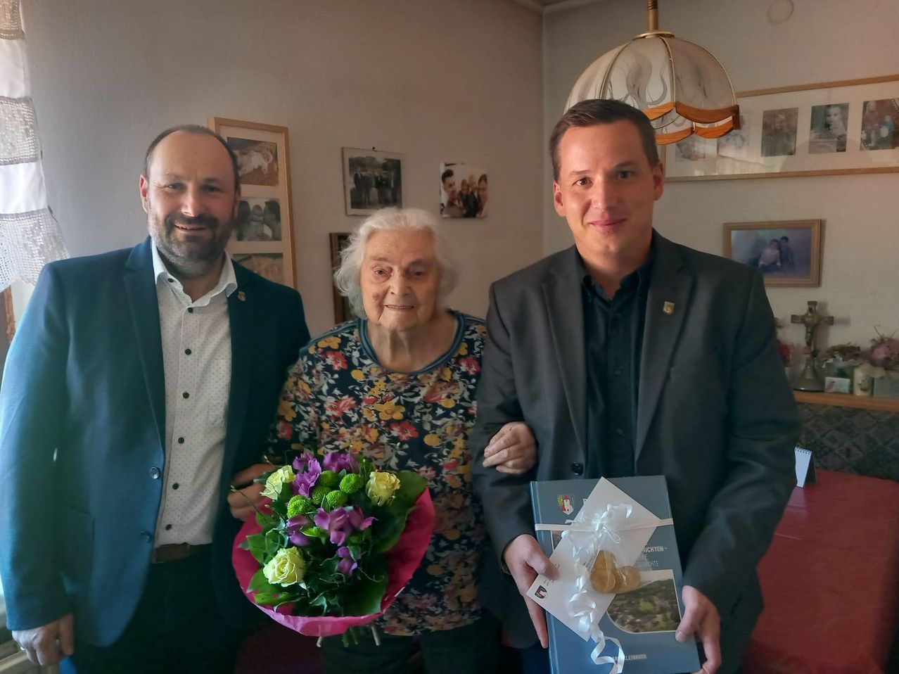 Drei Personen stehen gemeinsam in einem Raum. Die Frau hält einen Blumenstrauß, und der Mann ein Buch. Der Mann lächelt und die Frau schaut in die Kamera. Hinter ihnen zieren Familienfotos die Wand.