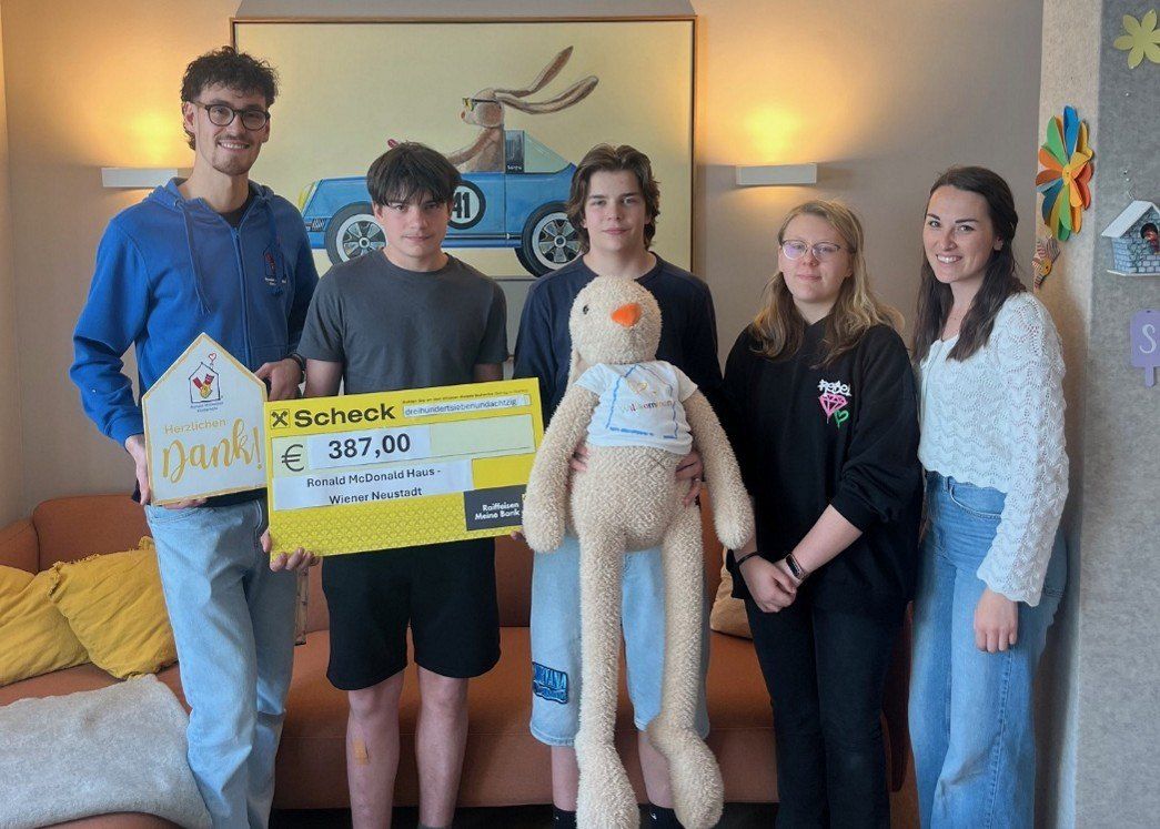 Eine Gruppe von fünf jungen Leuten steht drinnen mit einem Plüschtier und hält einen großen Scheck mit dem Betrag von 387,00 Euro. Sie lächeln und posieren für ein Foto.