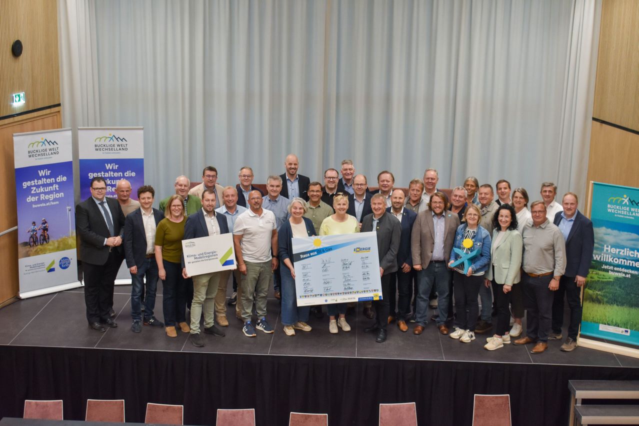Gruppenfoto von Menschen auf der Bühne, einige halten Urkunden. Eine Frau hält eine große Urkunde, andere kleinere. Ein Banner und Stühle im Hintergrund.
