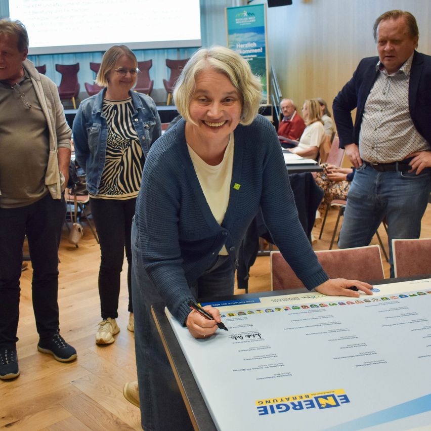 Eine Frau unterschreibt ein großes weißes Schild mit den Worten 'Energie Zukunft' in einem Raum, in dem andere Leute stehen.
