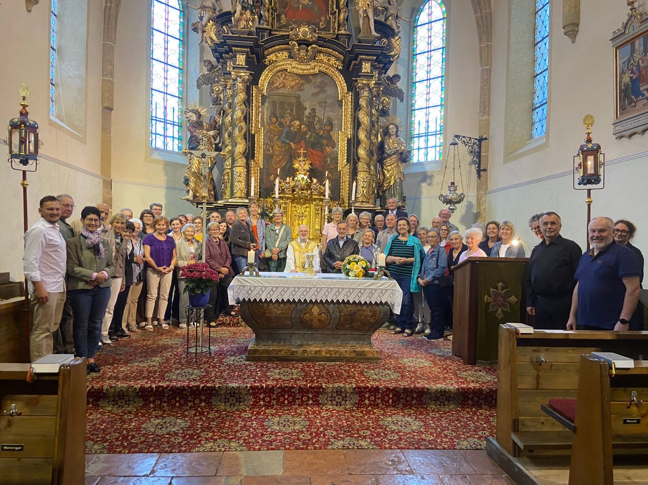 Eine Gruppe von Menschen steht vor einem Kirchenaltar mit Blumen und Kerzen, unter einem großen, kunstvollen Gemälde.