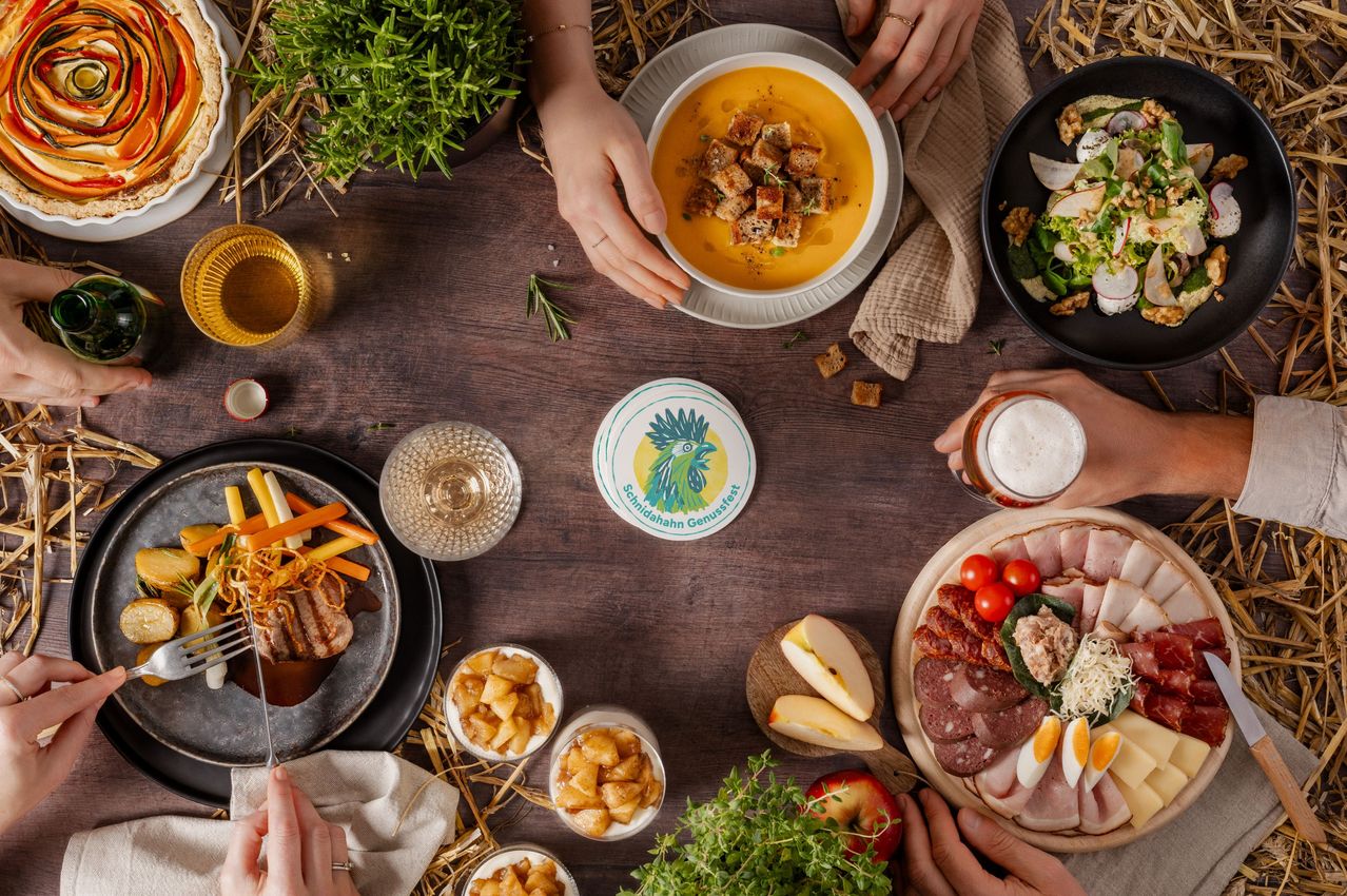 Ein Holztisch ist mit verschiedenen Gerichten gedeckt, einschließlich einer Suppenschüssel mit Croutons, einem Fleischteller, einem Salat und einem Glas Bier. Ein Logo für 'Schnabelahahn Genussfest' ist auf einem runden Teller zu sehen.