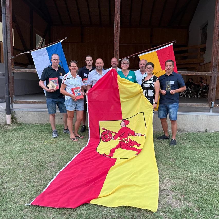 Eine Gruppe von Menschen steht vor einem Gebäude, hält eine Flagge und Bücher. Sie lächeln und posieren für ein Foto.