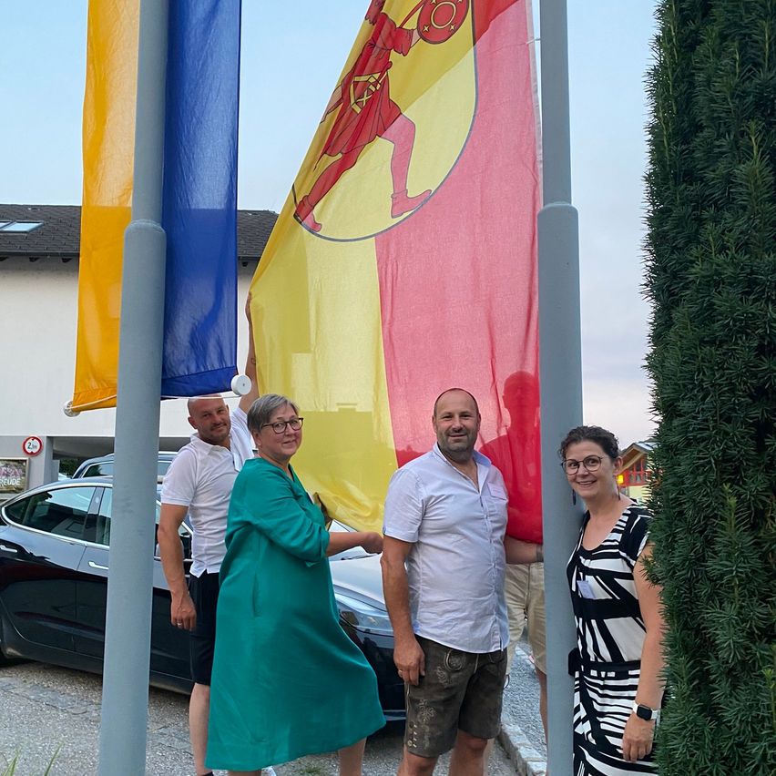 Vier Personen stehen neben einem Fahnenmast mit einer Fahne, die einen Ritter auf rotem Hintergrund zeigt, vor dem Hintergrund eines Gebäudes und eines Autos.