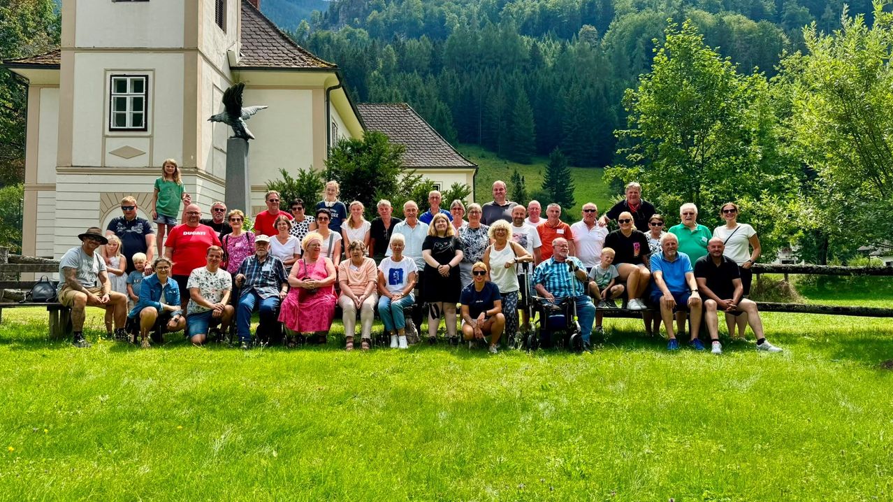 Eine Gruppe von Menschen, darunter einige im Rollstuhl, posiert für ein Foto vor einem Gebäude mit einer Statue und einem Berg im Hintergrund.