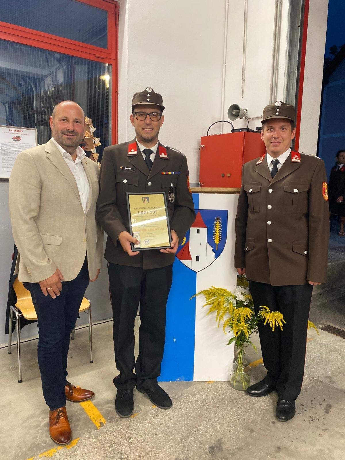 Drei Männer stehen zusammen, einer hält eine Plakette, während zwei andere in Uniformen auf beiden Seiten stehen. Dahinter befindet sich eine Wand mit einer Flagge und einer Pflanze in einer Vase.
