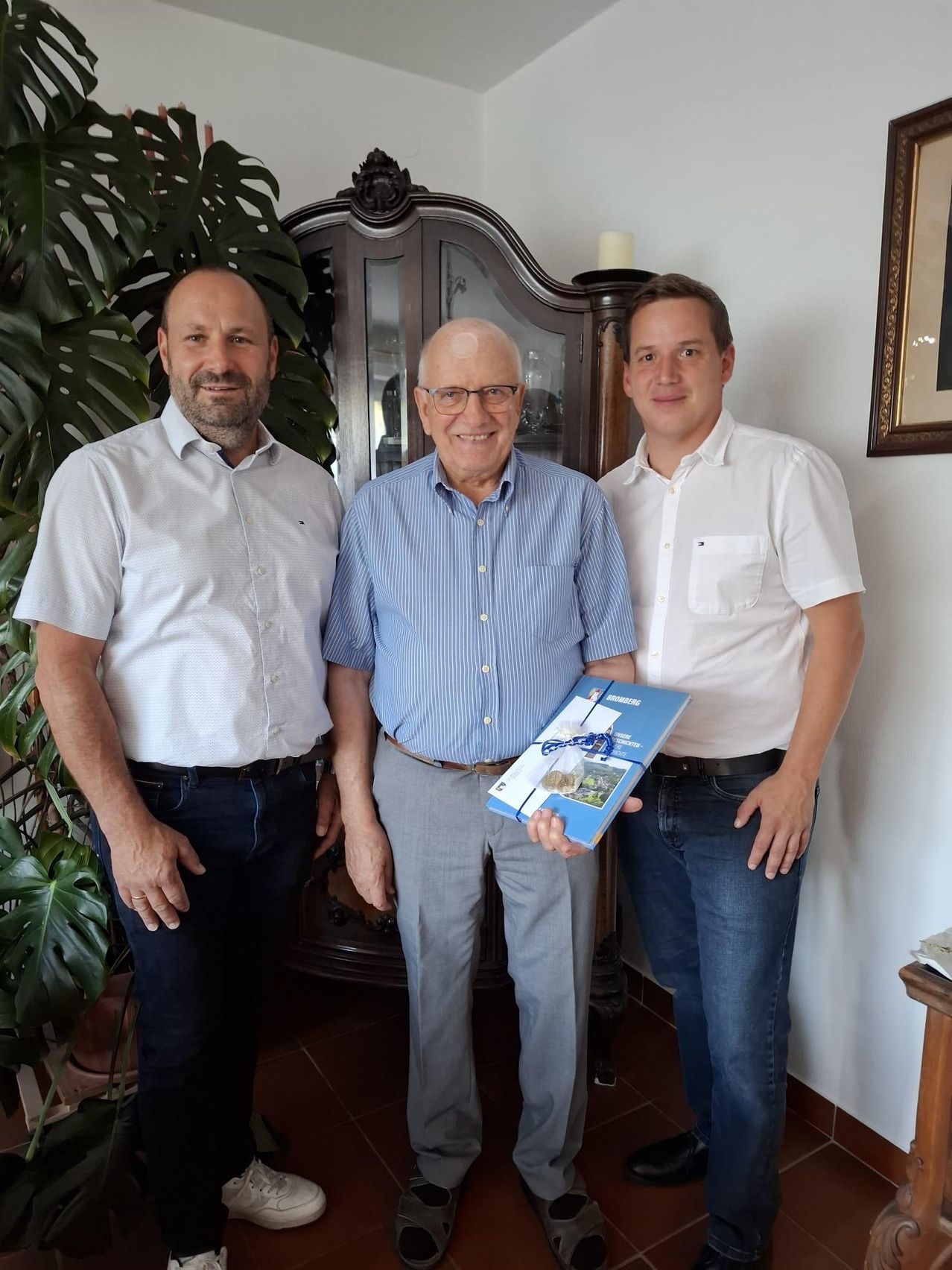 Three men stand together in a room, one holding a book. The man on the left wears a white shirt and jeans, the middle man wears a blue shirt and glasses, and the right man wears a white shirt and jeans. Behind them is a cabinet with a candle and a picture frame.