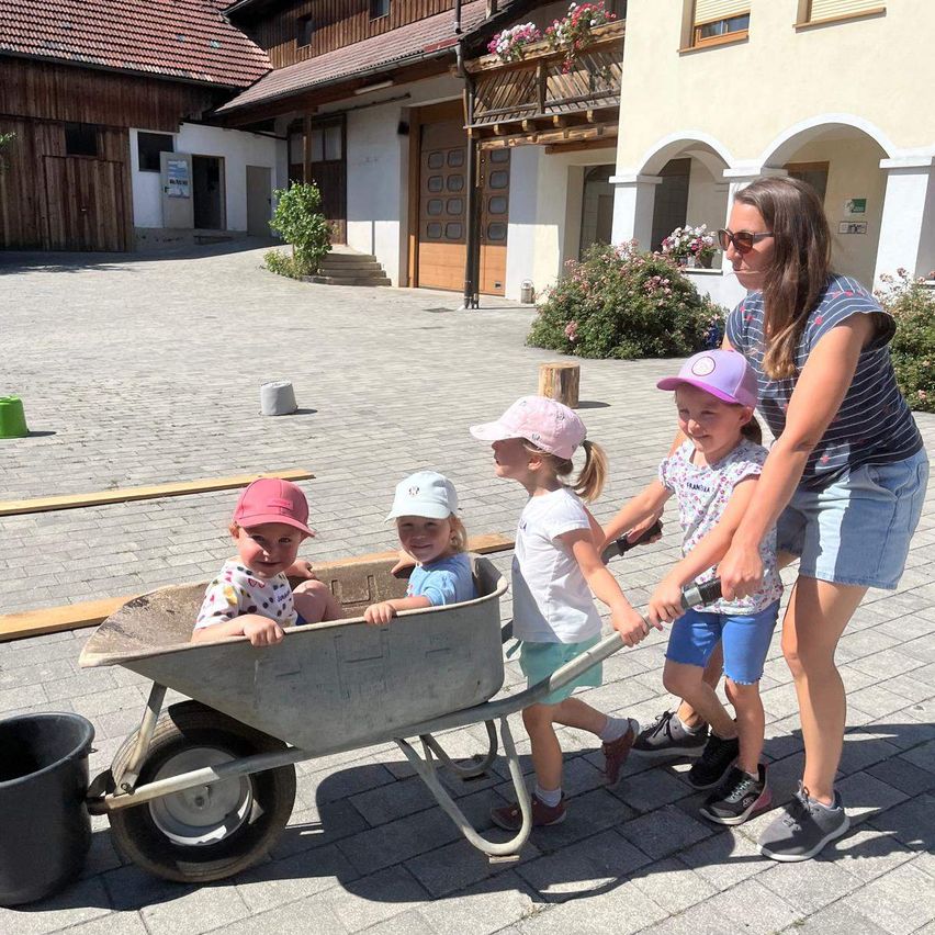 A woman and four children are pushing a wheelbarrow in a courtyard. The children wear hats and sneakers. Behind them, a building with a balcony and plants is visible.