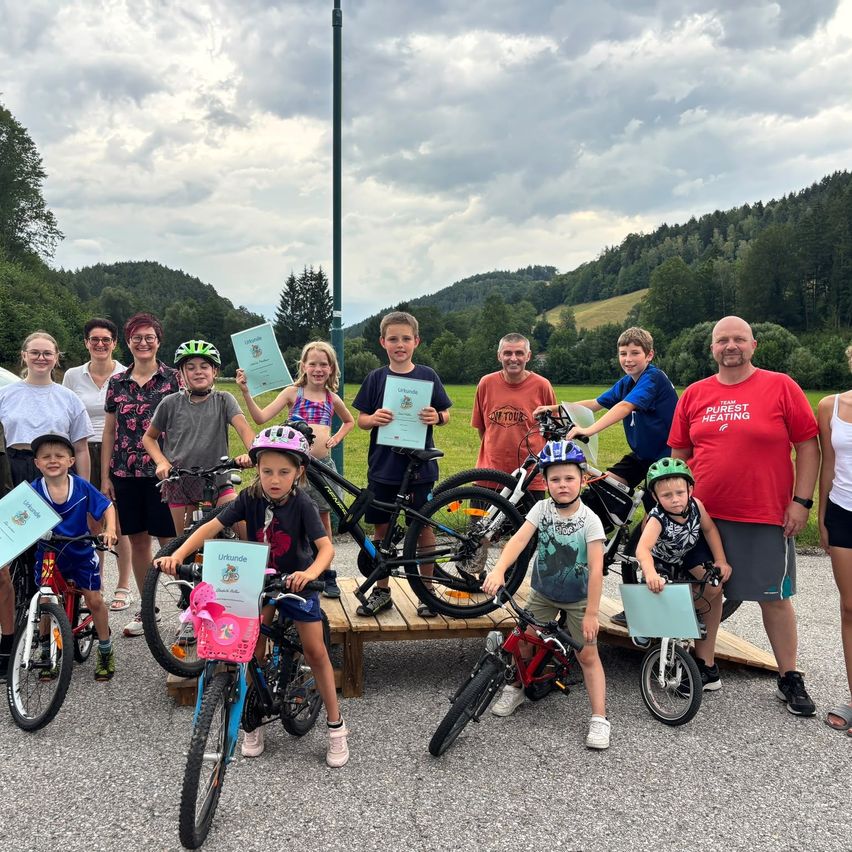 A group of children and adults pose for a photo with their bicycles and certificates in a scenic outdoor setting.