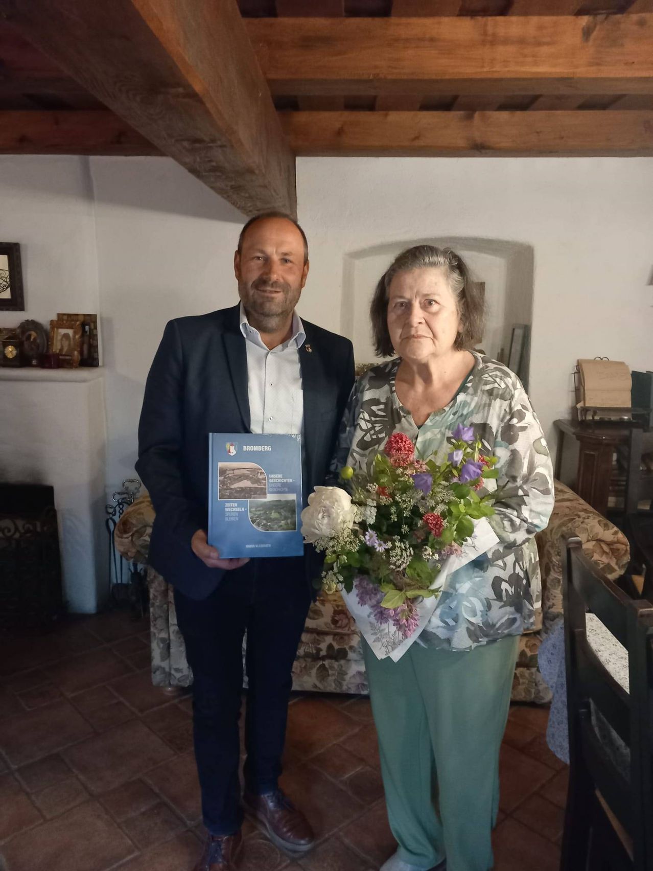 Ein Mann in einem Anzug hält ein Buch in der Hand und steht neben einer Frau mit einem Blumenstrauß. Sie befinden sich in einem Raum mit weißen Wänden, einem Kamin und einem Bücherregal.
