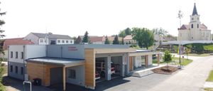 Eine moderne Feuerwache mit einem Feuerwehrauto in der Garage, umgeben von einem gepflasterten Parkplatz und Bäumen.