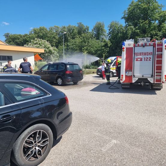 Ein Feuerwehrauto sprüht Wasser auf ein Gebäude. Ein Polizeiauto und ein Krankenwagen sind in der Nähe geparkt. Mehrere Menschen stehen draußen. Ein schwarzes Auto ist im Vordergrund geparkt.