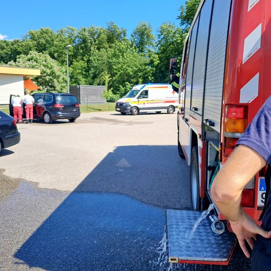 Ein Feuerwehrfahrzeug füllt an einem sonnigen Tag seinen Wassertank auf, mit einem geparkten Krankenwagen in der Nähe und zwei Personen, die bei einem Auto stehen.