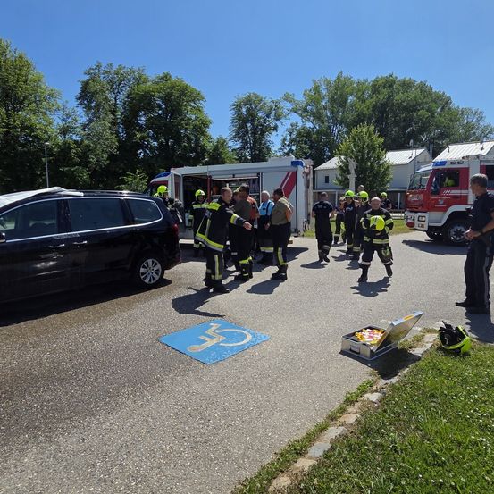 Eine Gruppe Feuerwehrleute versammelt sich auf einem Parkplatz mit zwei Feuerwehrfahrzeugen. Ein Mann steht rechts und ein Helm liegt auf dem Boden.