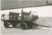 Ein altes Foto zeigt einen Traktor mit einem großen offenen Anhänger, der vor einem Gebäude geparkt ist.